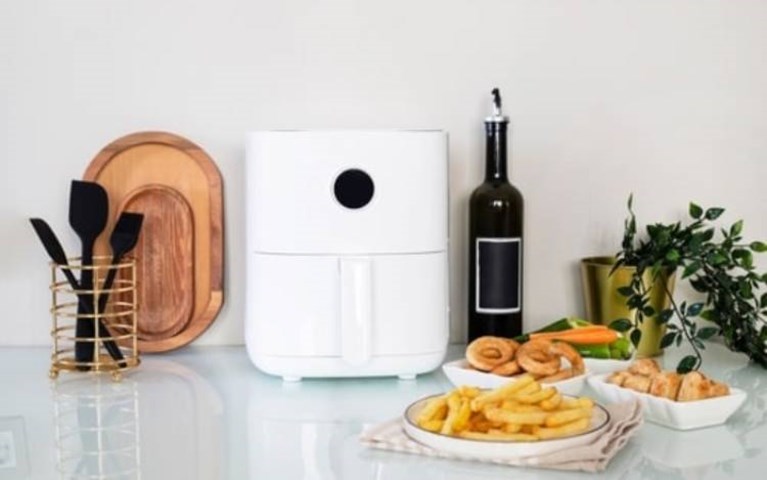 Clean white air fryer sat on a white reflective work surface with utensils and two wooden chopping boards to the left and olive oil, chips, onion rings, sausage rolls and a plant to the right.