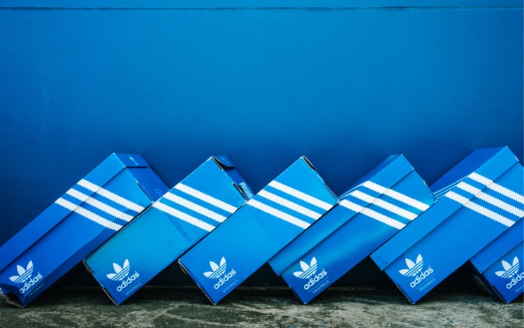 Six blue shoe boxes strewn on top of one another in a diagonal line, all of them have three white stripes on them and the Adidas logo on the bottom left.