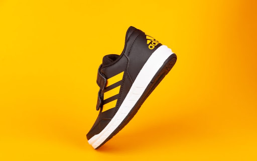 A clean black and yellow trainer with velcro straps and a white sole, facing sideways with three yellow vertical stripes on the side, photographed against a deep yellow background