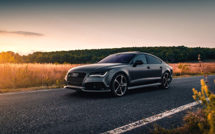 A black Audi alone on a road at sunset. The roadside is lined with brown grass/foliage and there are dark green trees in the background