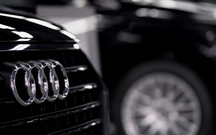 A black and white photograph of an Audi car grille and its silver logo featuring four rings. Another car and its front right wheel are partially visible, but blurred, in the background