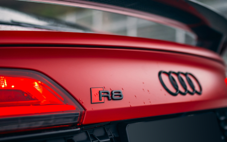 A close-up of the back of a red Audi racecar, with the black spoiler, black logo and left tail light visible