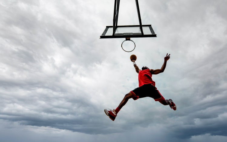 Low shot angle of a basketball player scoring a goal in the hoop with their legs spread in and arms wide, photographed against a cloudy sky