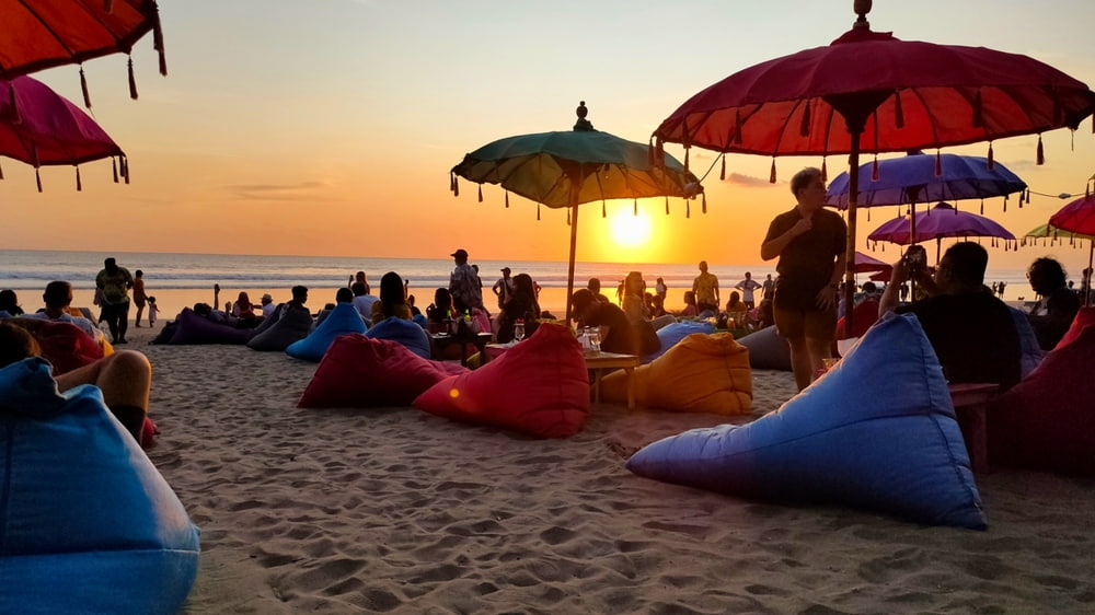 Tourists and locals enjoy the sunset over the famous Seminyak beach in Bali. People are relaxing on loungers under colourful beach umbrellas