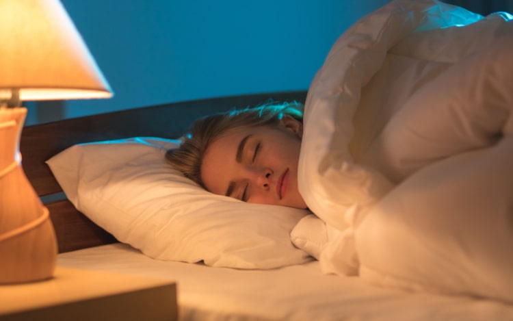 A white woman with blonde hair is asleep in bed, under a white duvet and on top of a white pillow. There is a lamp on the left-hand side of the image, turned on