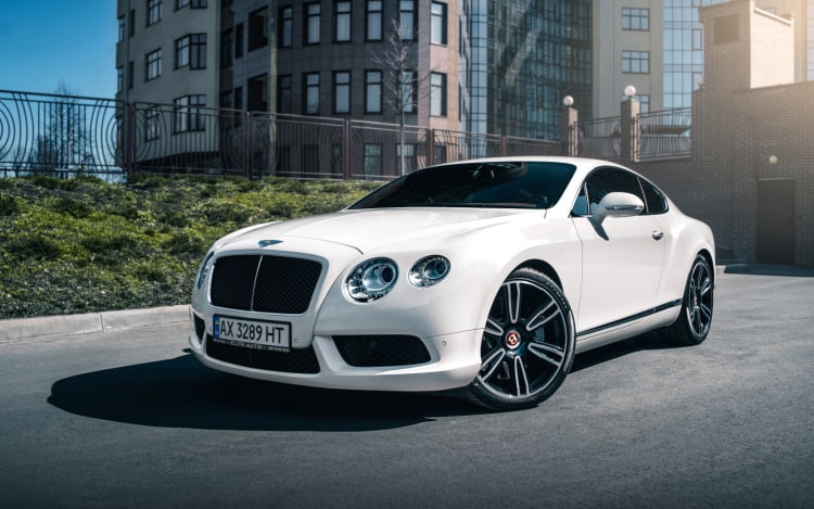 A white Bentley car with black wheels stationary on a grey road with a black fence and grass to the left and a partial view of tall buildings in the background.