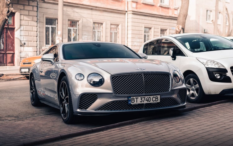 A silver Bentley parked at a kerb, with a white car parked next to it and stone-coloured buildings in the background.