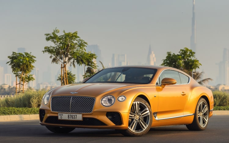 A gold-coloured Bentley stationary on a black road with small trees behind it and a city skyline in the distance.