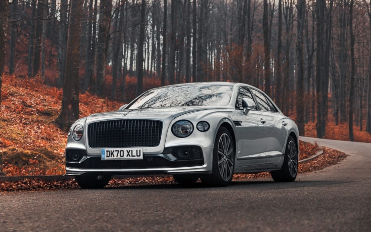 A silver Bentley on a winding grey road in a forest. There are fallen red leaves on the left and several tall thin trees behind the car.