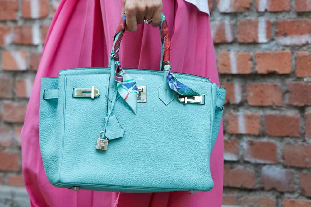 A person in a bright pink skirt stands next to a brick wall, holding a vivid turquoise leather Birkin bag.
