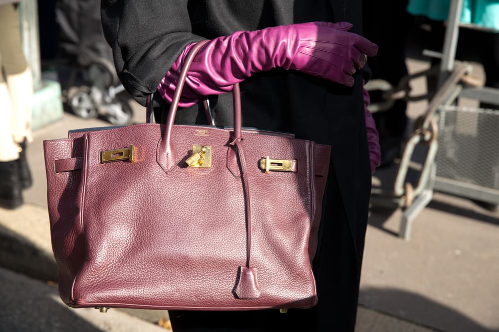 Some in a black coat and long purple gloves carrying a purple Birkin bag