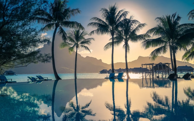 An infinity pool at sunset, with several palm trees reflected in the water. There are deck chairs and the silhouette of volcanic hills in the distance