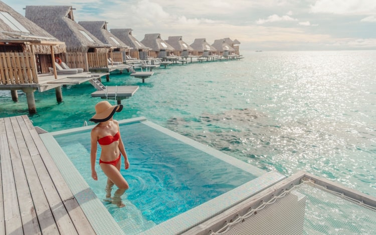 A woman in a red bikini and sun hat stepping into a pool next to the sea, with a line of beach huts next to her