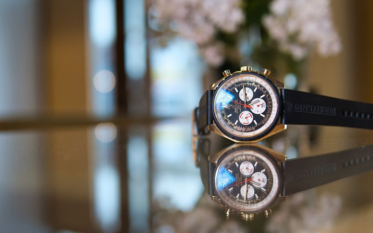 A navy Breitling watch with gold and white accents sitting on a reflective countertop with pale pink flowers and an open door out of focus in the background