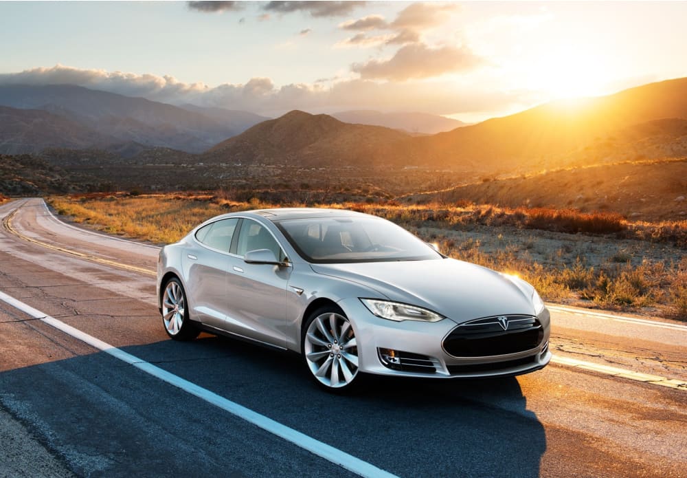 Grey sports car driving down a road with mountains in the background and no other cars in sight. To the top right of the image, the sun is setting over a mountain, and there are a few clouds.