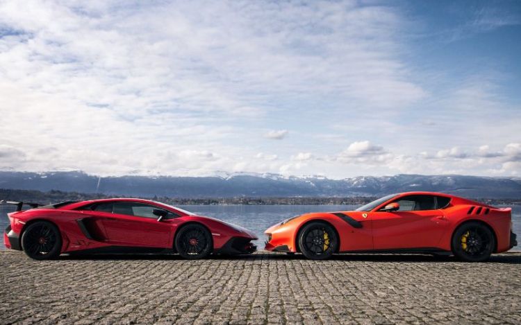 A red Lamborghini and orange Ferrari parked on a cobbled street by the water. The cars are facing each other, nearly touching