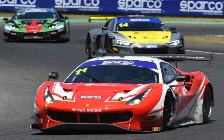 Three Ferrari racecars driving on a racetrack. A red Ferrari is in the foreground, followed by a silver Ferrari with a yellow bonnet, then a green and red Ferrari. The navy advertisement banner in the background says ‘sparco’ in white writing