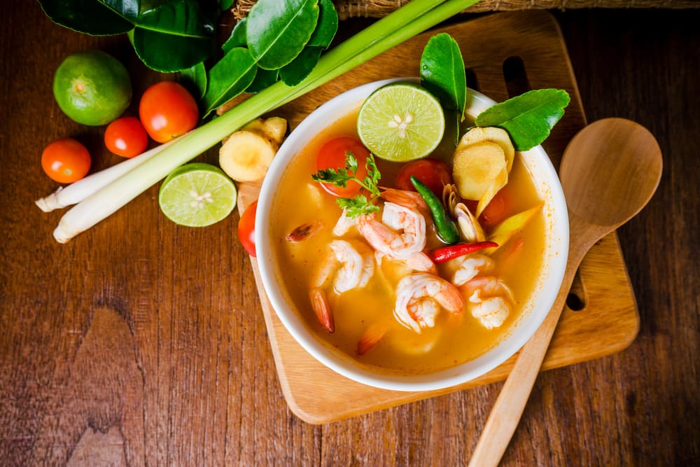 Tom yam kong, spicy clear orange soup with slices of lime on a wooden board next to some vegetables and a wooden spoon