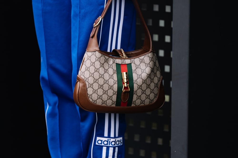 A close-up view of a person's legs wearing blue Adidas bottoms and holding a Gucci bag at knee length