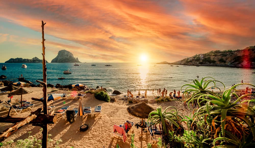 People hanging out at Cala d'Hort tropical beach with a sunset view of Es Vedra rock