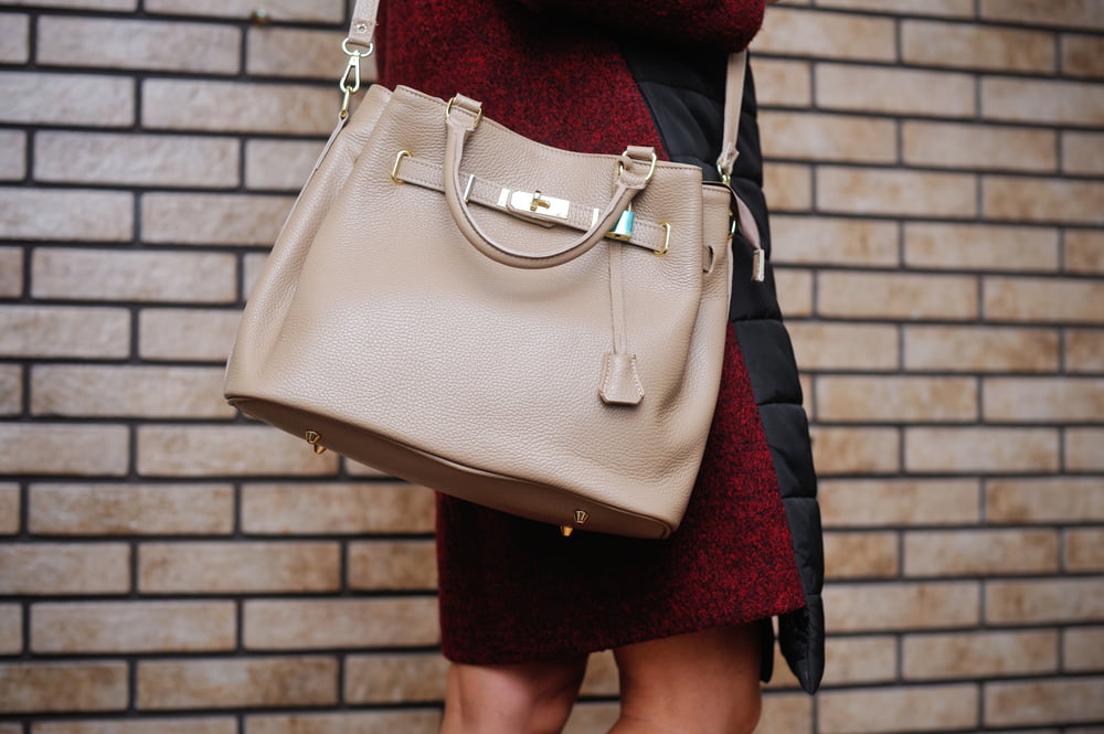 A person, wearing a red and black coat, walks next to a tiled wall. The person is holding a beige Kelly bag over their shoulder.