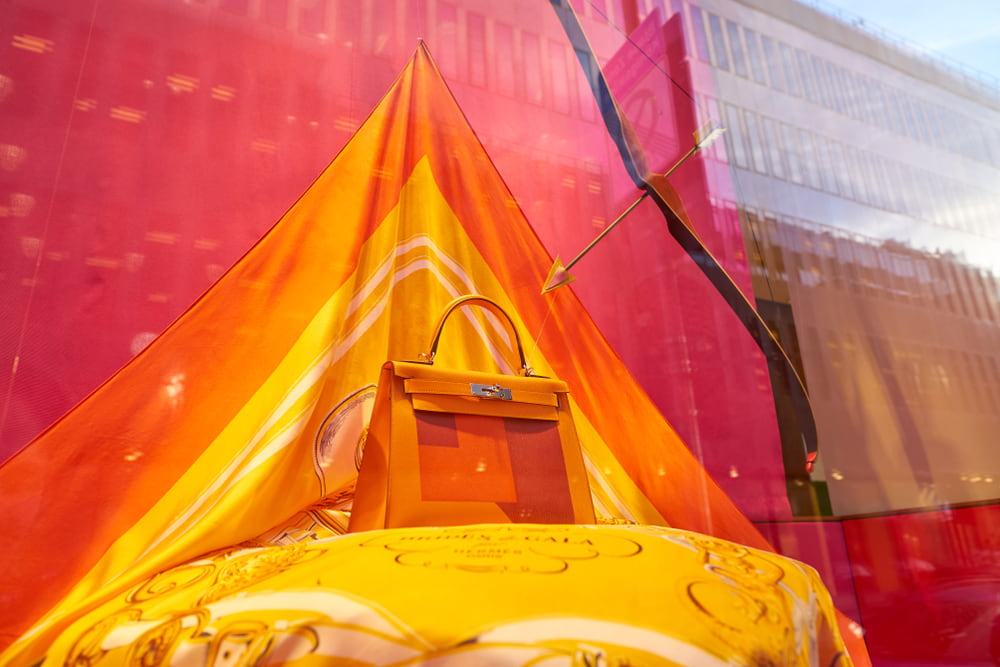 An orange Kelly bag on display in a storefront. The bag is sitting on an orange and yellow cloth, with pink wallpaper in the background.