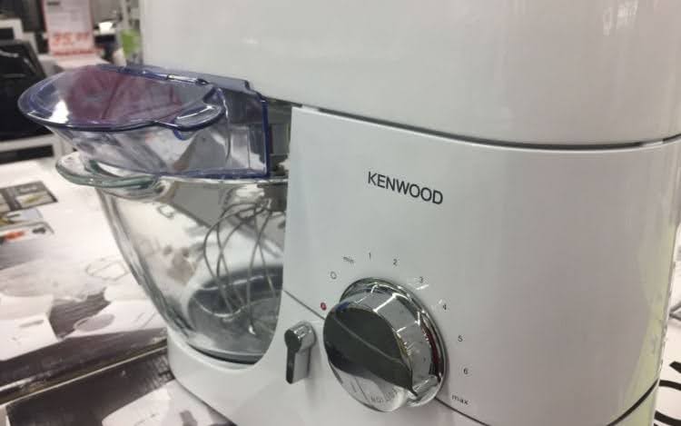 A white stand mixer with a silver Kenwood logo, a silver dial and a clear bowl attached.