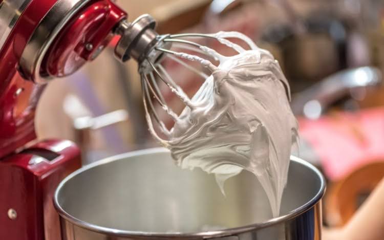 A red KitchenAid stand mixer with a white beaten mixture attached to the beater and a silver bowl partially in view beneath.