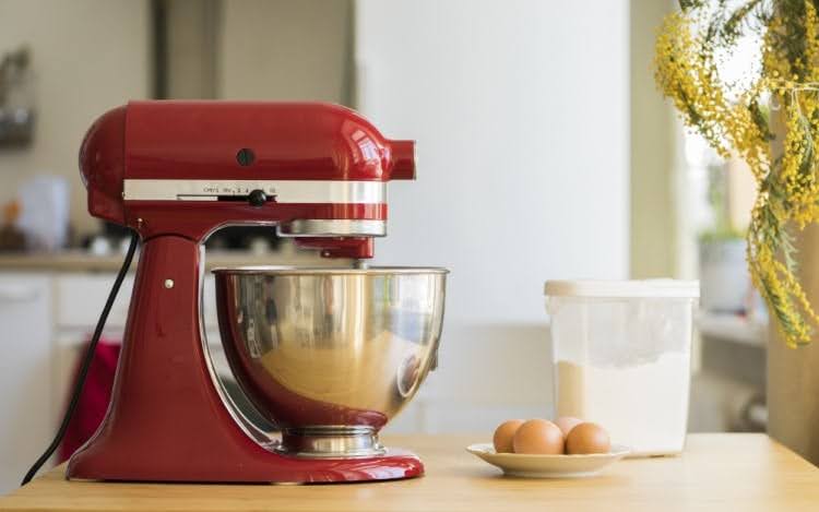 A red and silver KitchenAid stand mixer with a silver bowl attached. There are eggs on a plate and a clear container of flour next to the mixer.