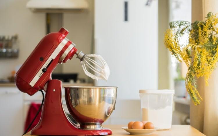 A red stand mixer with cream on the beater over a large silver bowl. There are eggs, a clear container of flour and yellow flowers to the right.