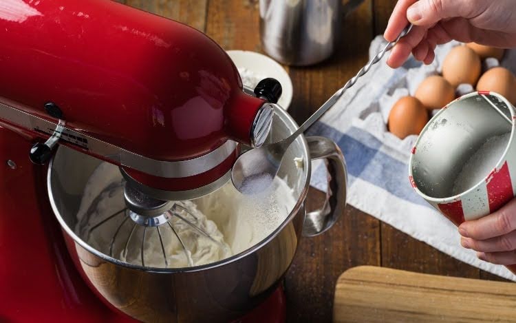 A red stand mixer beating a white substance in a silver bowl. Someone is adding sugar with a spoon, and there are eggs in the background.
