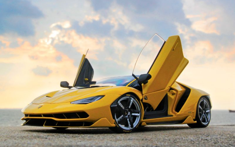 A yellow Lamborghini sports car under a pastel-coloured cloudy sky. The car is parked and both of its doors are opened upwards, the handles pointing towards the sky
