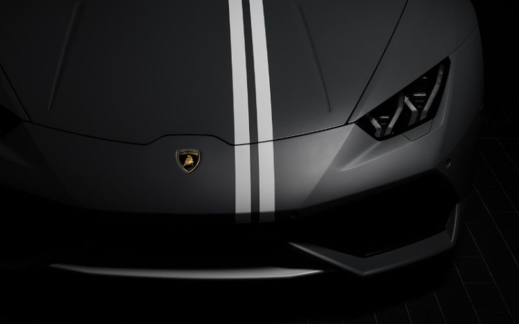 A matte grey Lamborghini showing only the bonnet and one headlight, with the other headlight in shadow. There are two white stripes running vertically from the grille up to the top of the image