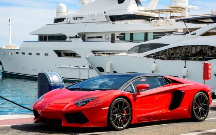 A red Lamborghini is parked on a dock with two large white yachts in the water behind it