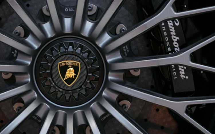 A close-up image of a car wheel, with the gold Lamborghini logo of a bull in the centre surrounded by silver spokes. Partially visible silver text of the brand name is on the right