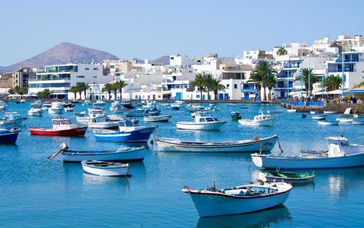 Several small boats floating on calm blue waters, with a row of white houses and buildings in the background and a mountain in the distance