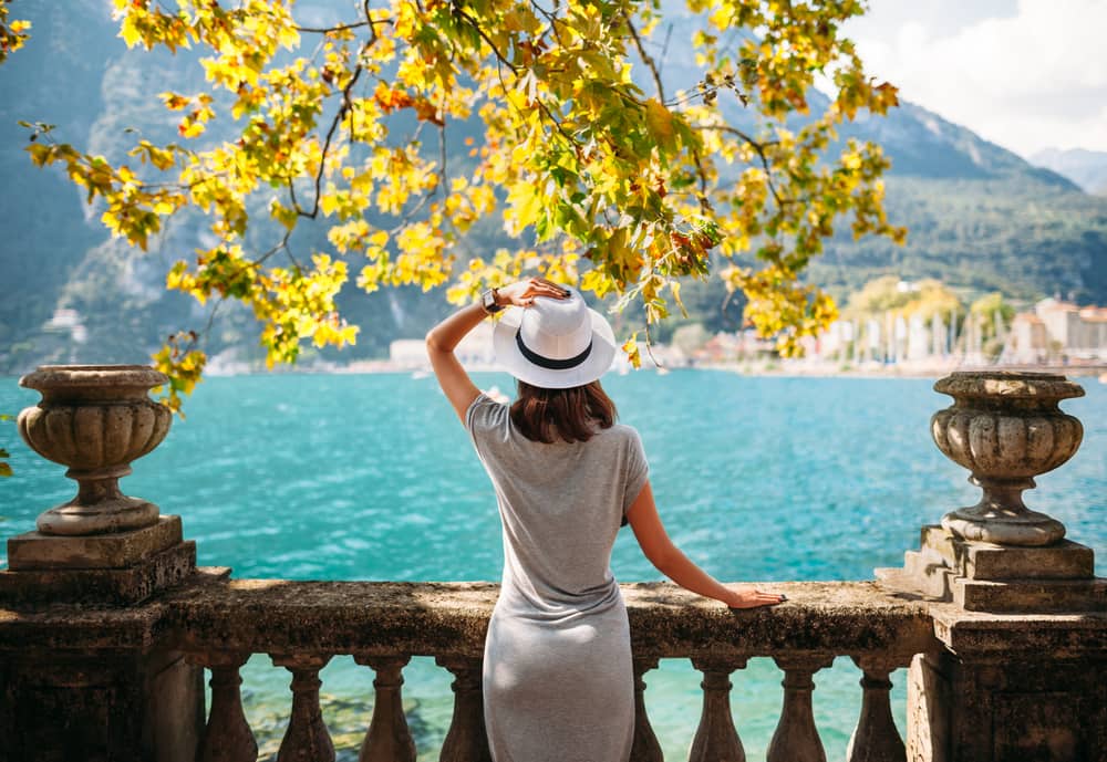 Person holding onto their white hat while they look at the views of Lake Garda as they stand against an old-fashioned stone barrier underneath branches with yellow leaves