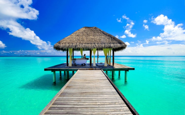 A wooden walkway heading out to sea with a beach hut at the end. The water is turquoise and the sky is blue with a few white clouds