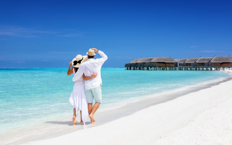 A couple in white walk along a white sand beach next to a clear blue sea. There are beach huts in the water in the background