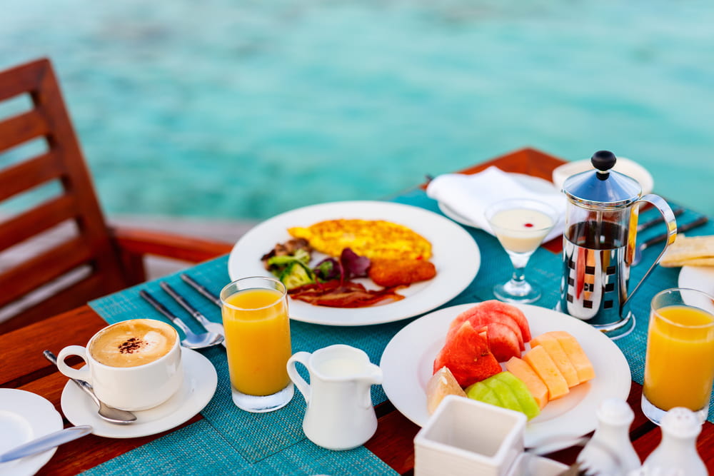 Table corner with a plate of fruit, a plate with breakfast, two glass of orange juice, a cup of coffee and a cafetiere with blue water blurred in the background