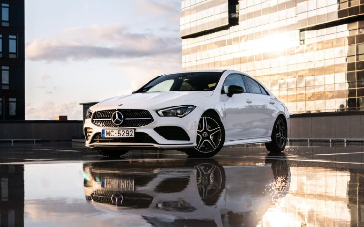 A white Audi parked on a rooftop car park with a glass skyscraper in full sunlight behind it and a puddle reflecting both the car and part of the building