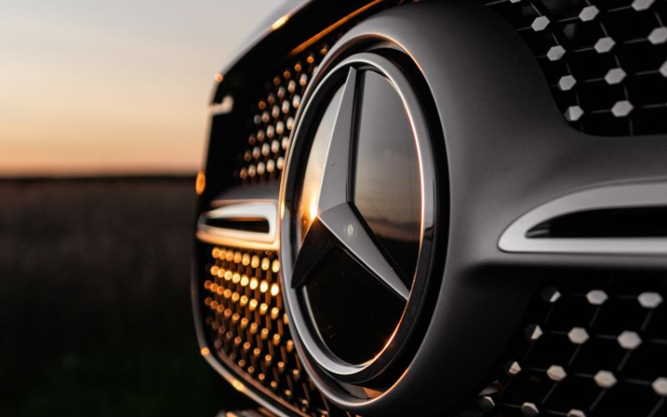 A close-up shot of the silver and black Mercedes logo on the front of a black car. Part of the grille is slightly orange as it’s pointed towards the evening sun out of shot