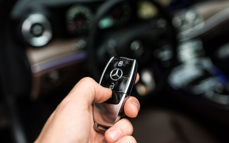 A white person’s hand holding a black Mercedes car key with their hand hovering over the unlock button. The interior of the car is blurred but visible in the background, including some of the dashboard and steering wheel