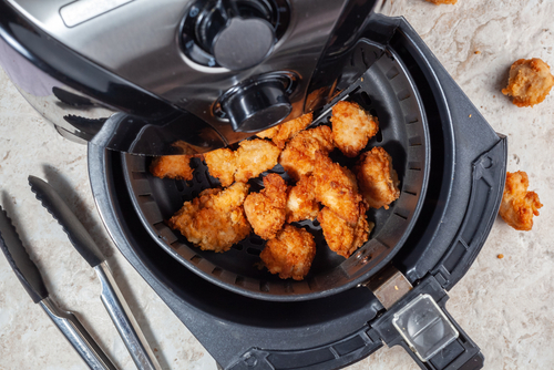 Silver air fryer with crispy chicken inside the drawer, set against a marble worktop with tongs to the left.