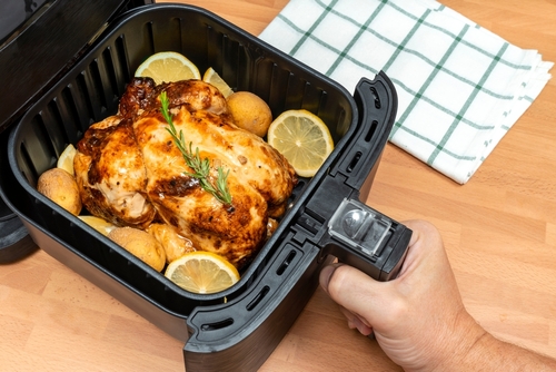 Black air fryer drawer with a whole roasted chicken inside with lemon slices, rosemary and potatoes. Being operated by a hand, set against a wooden work surface with a tea towel in frame.