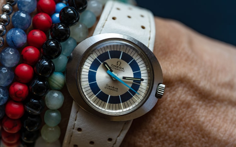An Omega watch on a woman’s wrist next to several beaded bracelets. The watch has a white leather strap, and a silver, white and navy blue face