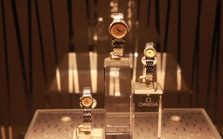 3 silver Omega watches on clear plastic stands with a brown background