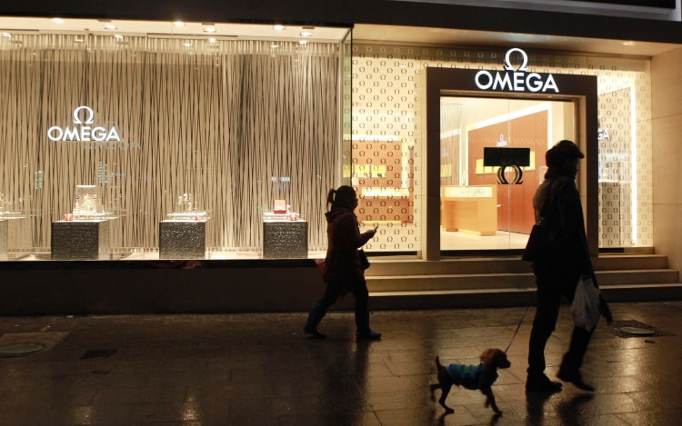  An Omega storefront at night. The lights are on inside. Two women are walking by. One woman has a dog on a lead