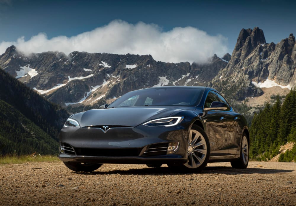 Front shot of a dark blue luxury 5 door sports car with modern features, on a gravel path with green mountains in the background in a partially cloudy setting.