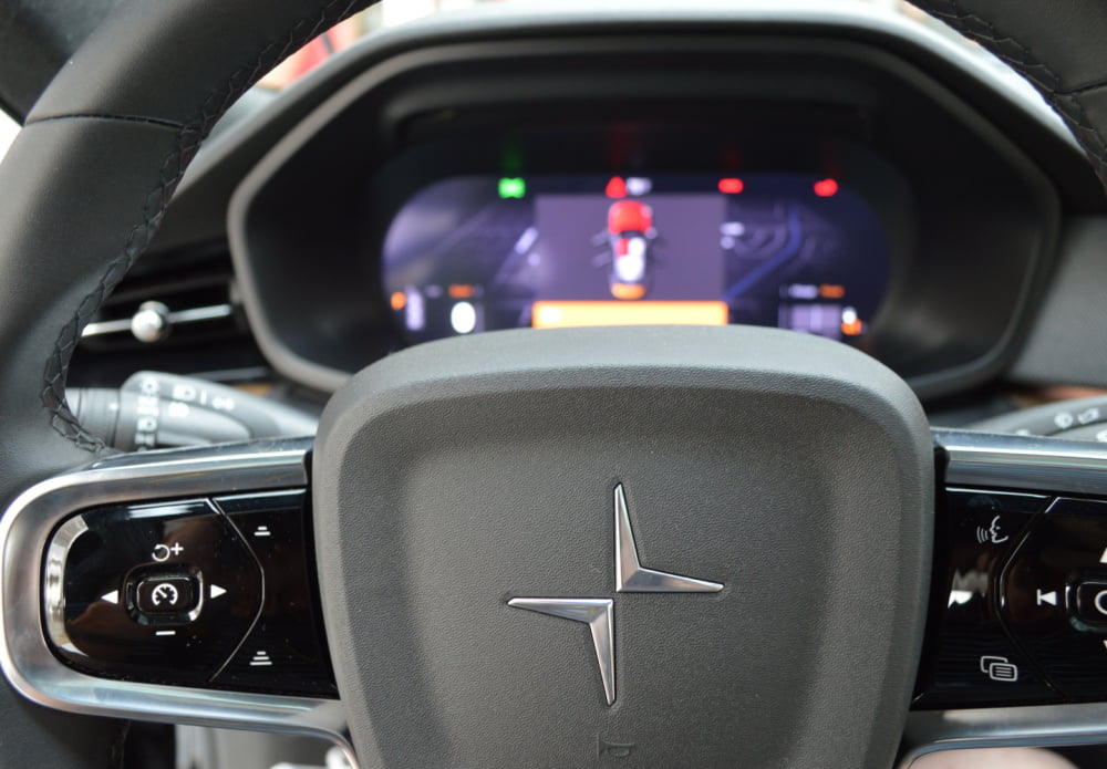 Close up of a car steering wheel with a dashboard in the background. To the sides of the centre of the steering wheel are buttons to control various elements of the car.